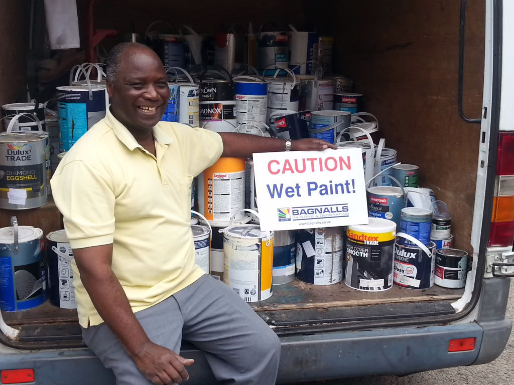 A Man Sits Next To Multiple Tins Of Paint In The Back Of A Van Holding A Wet Paint Sign