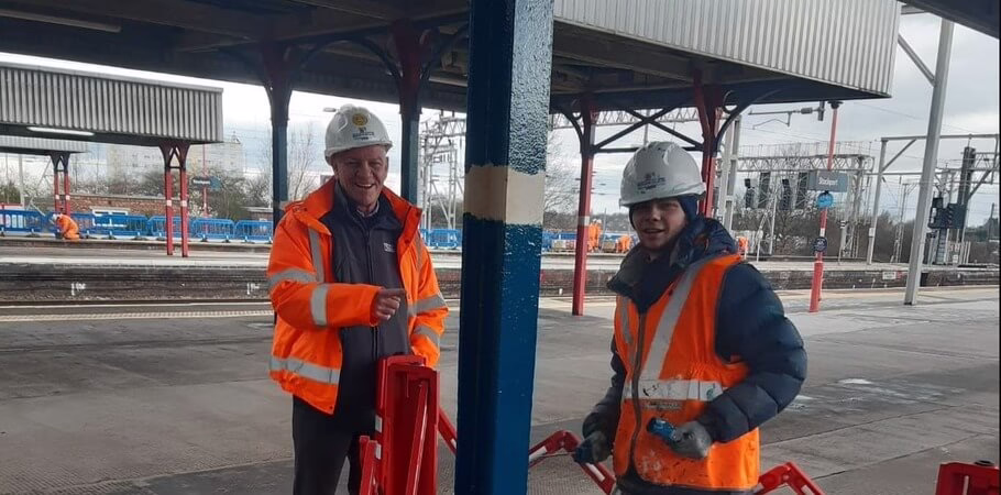 Two Bagnalls Team Members In Safety Gear Paint A Pillar Blue On A Train Station Platform