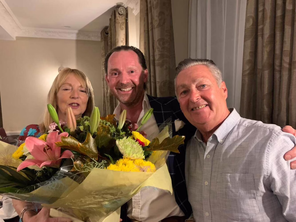 Stephen Bagnall Stands With Steve Bethell And Isobel Holding A Bunch Of Flowers