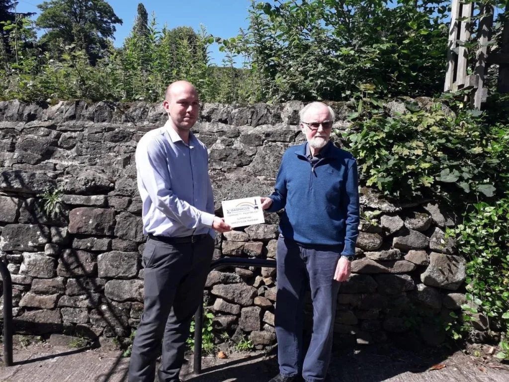Two Men Stand Outside In Front Of A Stone Wall Holding A Plaque Celebrating Bagnalls Contribution To The Shipley Glen Tramway